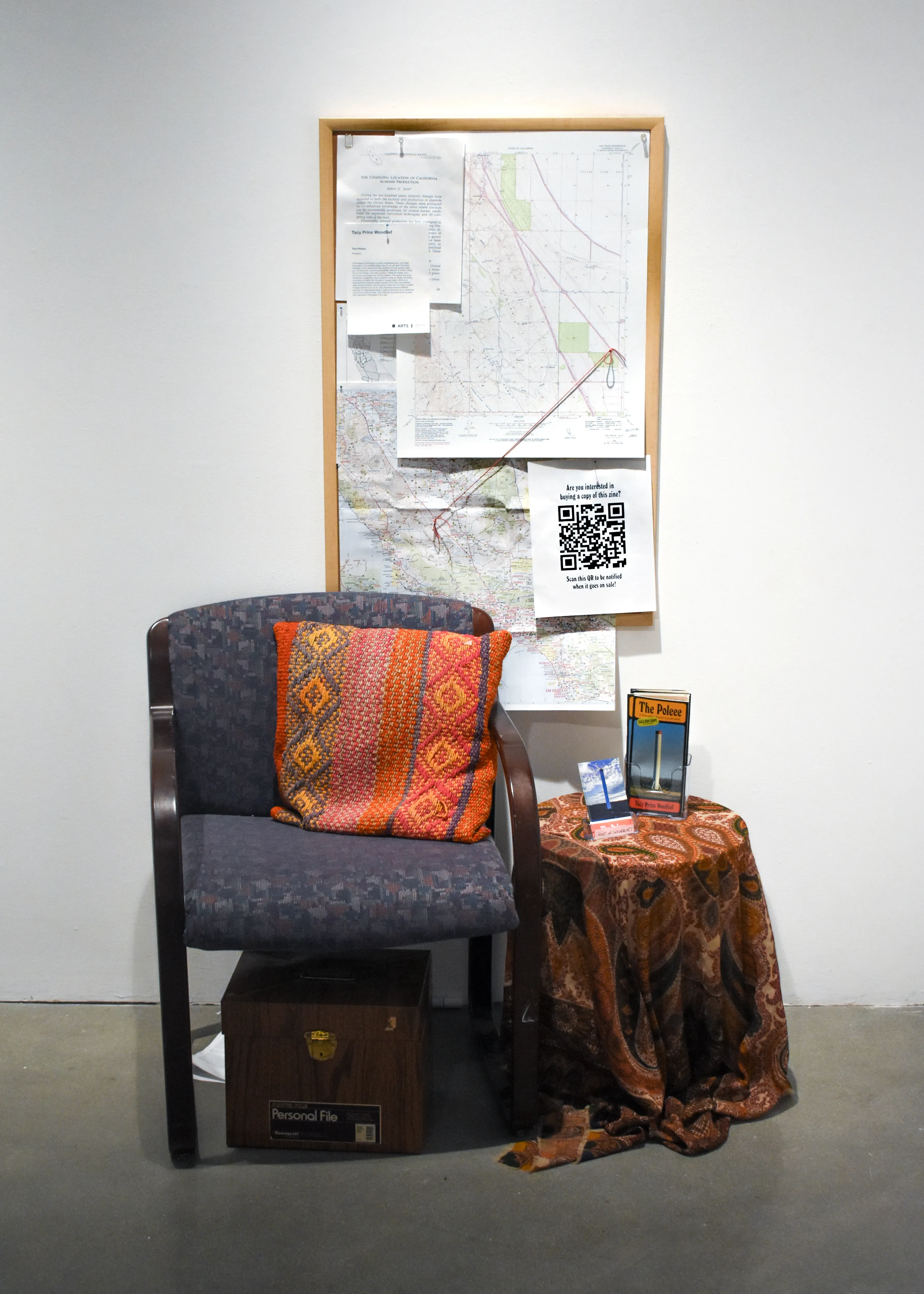 the interactive component of the installation, a chair sits in front of a bulletin board with maps pinned to it, and beside a table with a brochure holder holding copies of the zine.